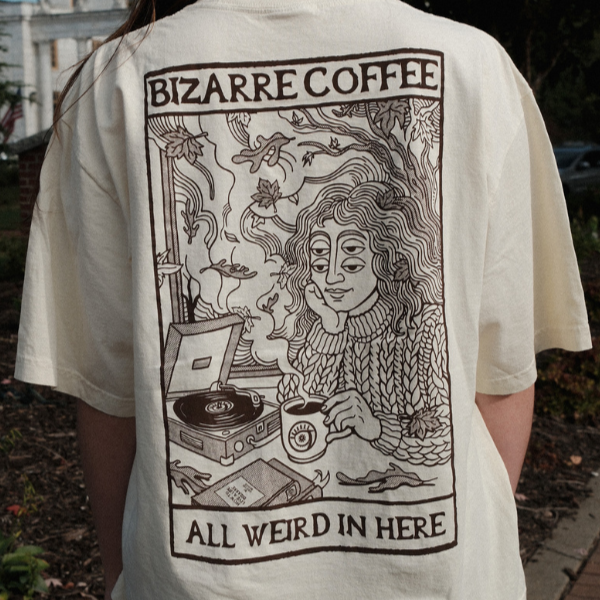 Beige t-shirt with a graphic design of a person with a record player and text 'Bizarre Coffee All Weird In Here' on the back.